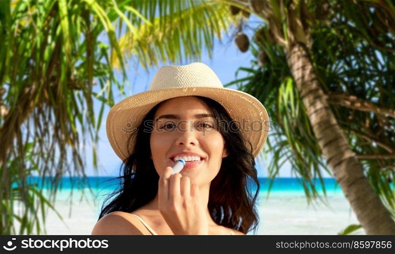 beauty, cosmetics and summer holidays concept - portrait of smiling young woman in bikini swimsuit and straw hat applying lip balm over tropical beach and palm trees background in french polynesia. smiling woman in bikini with lip balm on beach