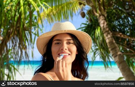 beauty, cosmetics and summer holidays concept - portrait of smiling young woman in bikini swimsuit and straw hat applying lip balm over tropical beach and palm trees background in french polynesia. smiling woman in bikini with lip balm on beach