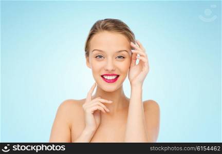 beauty, cosmetics, people and health concept - smiling young woman with pink lipstick on lips touching her face over blue background