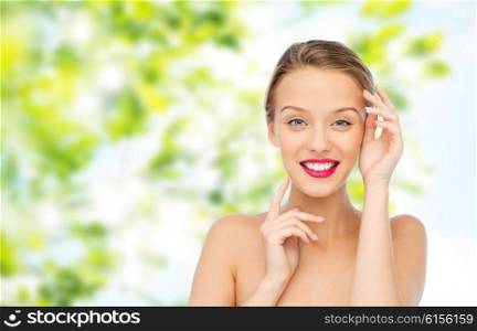 beauty, cosmetics, people and health concept - smiling young woman with pink lipstick on lips touching her face over green natural background