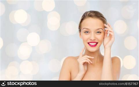 beauty, cosmetics, people and health concept - smiling young woman with pink lipstick on lips touching her face over holidays lights background