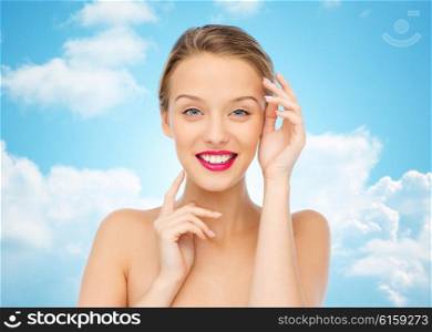 beauty, cosmetics, people and health concept - smiling young woman with pink lipstick on lips touching her face over blue sky and clouds background