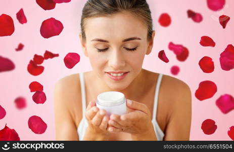 beauty, cosmetics, skin care and people concept - close up of beautiful happy woman holding and smelling cream jar over red rose petals on pink background. close up of woman smelling cream over rose petals. close up of woman smelling cream over rose petals