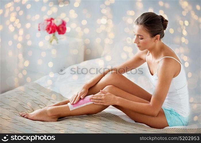 beauty, depilation, epilation, hair removal and people concept - beautiful woman applying depilatory wax strip to her leg skin at home bedroom. woman epilating leg hair with wax strip at home