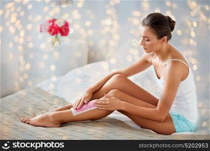 beauty, depilation, epilation, hair removal and people concept - beautiful woman applying depilatory wax strip to her leg skin at home bedroom. woman epilating leg hair with wax strip at home