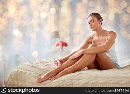 beauty, epilation and bodycare concept - beautiful woman with epilator removing hair from legs sitting on bed at home bedroom. woman with epilator removing hair on legs at home