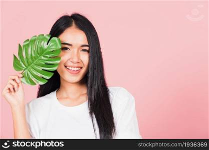 Beauty face. Portrait of Asian beautiful young woman with fresh healthy skin hold green monstera leaf on her face, Tropical Leaves, studio shot isolated on pink background, Skin body care spa concept