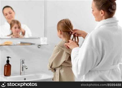 beauty, family and people concept - happy smiling mother and daughter looking to mirror and braiding hair in bathroom. mother and daughter braiding hair in bathroom