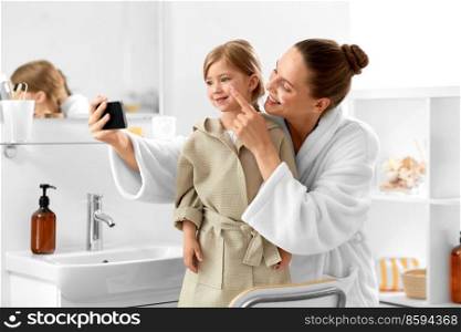 beauty, family and people concept - happy smiling mother and daughter with moisturizer on their faces taking selfie with smartphone in bathroom. mother and daughter taking selfie with in bathroom