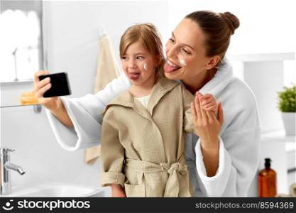 beauty, family and people concept - happy smiling mother and daughter with moisturizer on their faces sticking tongues and taking selfie with smartphone in bathroom. mother and daughter taking selfie with in bathroom