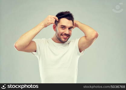 beauty, grooming and people concept - smiling young man brushing hair with comb over gray background. happy man brushing hair with comb over gray