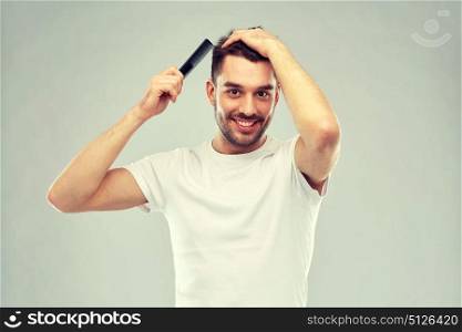 beauty, grooming and people concept - smiling young man brushing hair with comb over gray background. happy man brushing hair with comb over gray