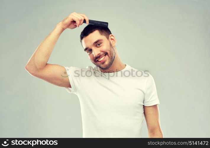 beauty, grooming and people concept - smiling young man brushing hair with comb over gray background. happy man brushing hair with comb over gray