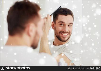 beauty, grooming and people concept - smiling young man looking to mirror and brushing hair with comb at home bathroom over snow