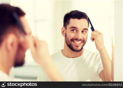 beauty, grooming and people concept - smiling young man looking to mirror and brushing hair with comb at home bathroom. happy man brushing hair with comb at bathroom