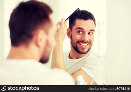 beauty, grooming and people concept - smiling young man looking to mirror and brushing hair with comb at home bathroom. happy man brushing hair with comb at bathroom