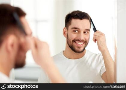 beauty, grooming and people concept - smiling young man looking to mirror and brushing hair with comb at home bathroom