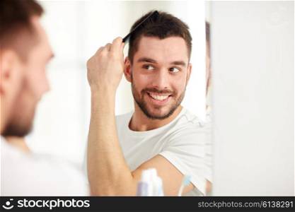 beauty, grooming and people concept - smiling young man looking to mirror and brushing hair with comb at home bathroom