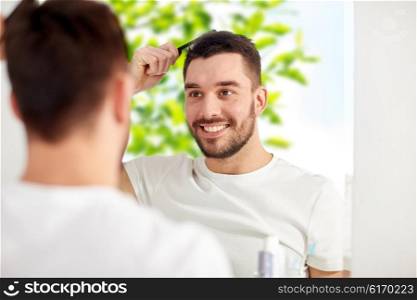 beauty, grooming and people concept - smiling young man looking to mirror and brushing hair with comb at home bathroom over green natural background