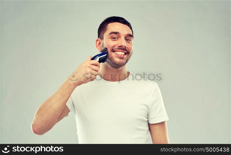 beauty, grooming and people concept - smiling young man shaving beard with trimmer or electric shaver over gray background. smiling man shaving beard with trimmer over gray