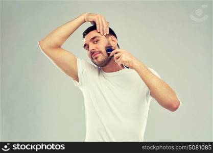 beauty, grooming and people concept - smiling young man shaving beard with trimmer or electric shaver over gray background. smiling man shaving beard with trimmer over gray