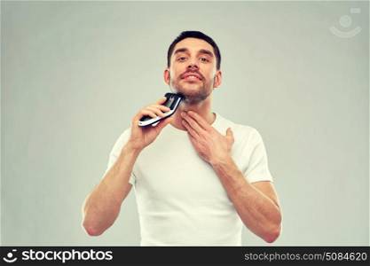 beauty, grooming and people concept - smiling young man shaving beard with trimmer or electric shaver over gray background. smiling man shaving beard with trimmer over gray. smiling man shaving beard with trimmer over gray