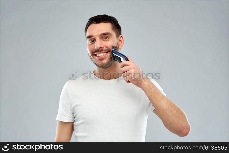 beauty, grooming and people concept - smiling young man shaving beard with trimmer or electric shaver over gray background