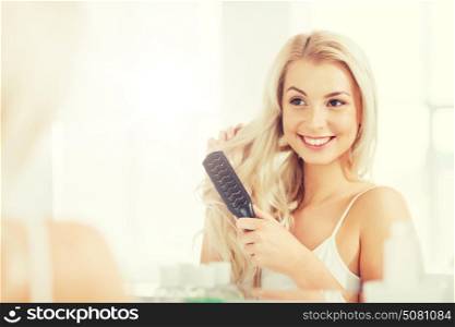 beauty, grooming and people concept - smiling young woman looking to mirror and brushing hair with comb at home bathroom. happy woman brushing hair with comb at bathroom