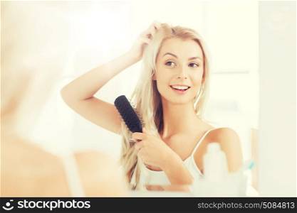 beauty, grooming and people concept - smiling young woman looking to mirror and brushing hair with comb at home bathroom. happy woman brushing hair with comb at bathroom. happy woman brushing hair with comb at bathroom