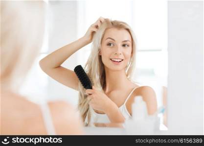 beauty, grooming and people concept - smiling young woman looking to mirror and brushing hair with comb at home bathroom