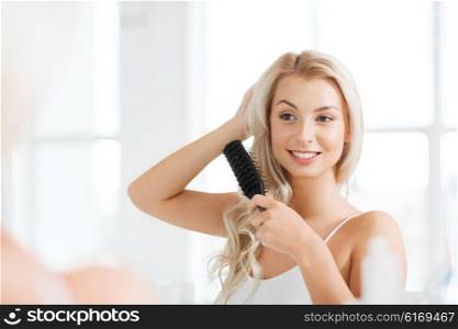 beauty, grooming and people concept - smiling young woman looking to mirror and brushing hair with comb at home bathroom