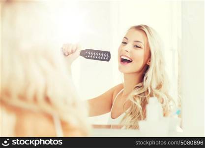 beauty, grooming, hair care and people concept - smiling young woman looking to mirror and singing to hair brush or comb at home bathroom. happy woman singing to hair brush at bathroom