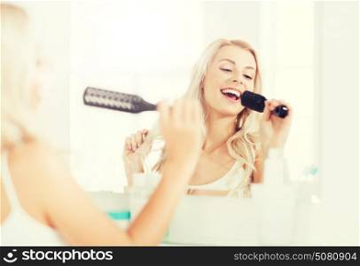 beauty, grooming, hair care and people concept - smiling young woman looking to mirror and singing to hair brush or comb at home bathroom. happy woman singing to hair brush at bathroom