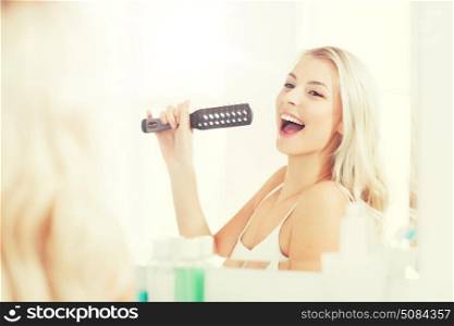 beauty, grooming, hair care and people concept - smiling young woman looking to mirror and singing to hair brush or comb at home bathroom. happy woman singing to hair brush at bathroom. happy woman singing to hair brush at bathroom