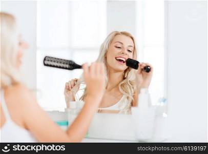 beauty, grooming, hair care and people concept - smiling young woman looking to mirror and singing to hair brush or comb at home bathroom