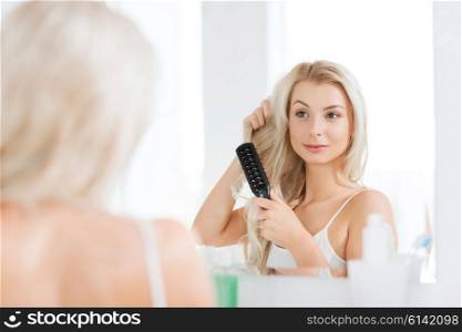 beauty, grooming, hair care and people concept - smiling young woman looking to mirror and brushing hair with comb at home bathroom