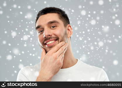 beauty, grooming, winter, christmas and people concept - happy young man touching his face or beard over snow on gray background