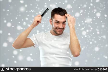 beauty, grooming, winter, christmas and people concept - smiling young man brushing hair with comb over snow on gray background