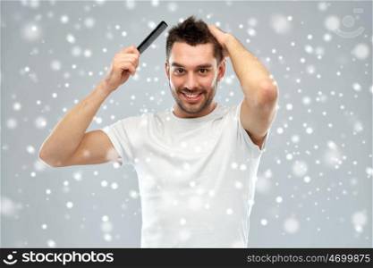 beauty, grooming, winter, christmas and people concept - smiling young man brushing hair with comb over snow on gray background. happy man brushing hair with comb over snow