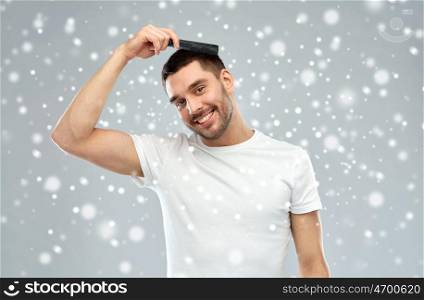 beauty, grooming, winter, christmas and people concept - smiling young man brushing hair with comb over snow on gray background