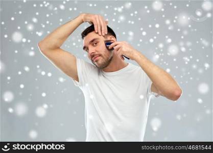 beauty, grooming, winter, christmas and people concept - smiling young man shaving whisker with trimmer or electric shaver over snow on gray background
