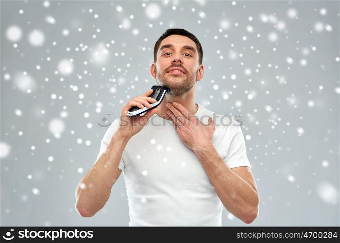 beauty, grooming, winter, christmas and people concept - smiling young man shaving beard with trimmer or electric shaver over snow on gray background