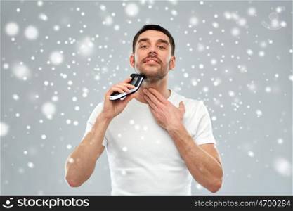 beauty, grooming, winter, christmas and people concept - smiling young man shaving beard with trimmer or electric shaver over snow on gray background