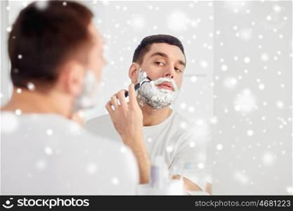 beauty, grooming, winter, christmas and people concept - young man looking to mirror and shaving beard with manual razor blade at home bathroom over snow