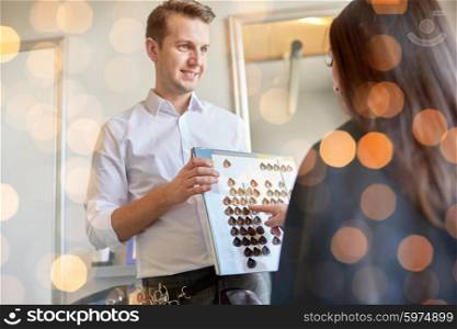 beauty, hair dyeing and people concept - happy young woman with hairdresser choosing hair color from palette samples at salon over holidays lights