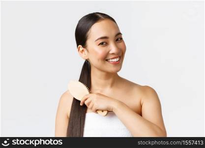 Beauty, hair loss products, shampoo and hair care concept. Close-up of beautiful asian female in bath towel brushing hair with brush and smiling, standing in bathroom over white background.. Beauty, hair loss products, shampoo and hair care concept. Close-up of beautiful asian female in bath towel brushing hair with brush and smiling, standing in bathroom over white background