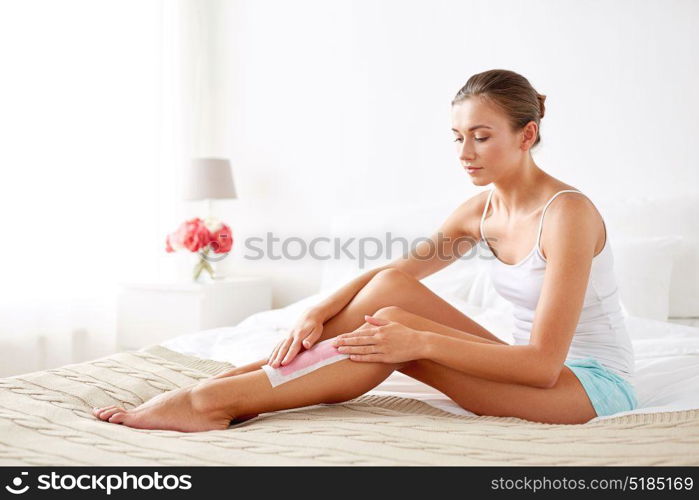 beauty, hair removal and people concept - beautiful woman applying depilatory wax strip to her leg skin at home bedroom. woman removing leg hair with depilatory wax strip