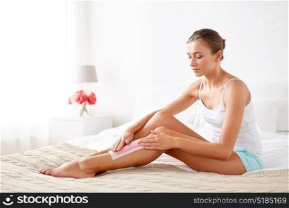 beauty, hair removal and people concept - beautiful woman applying depilatory wax strip to her leg skin at home bedroom. woman removing leg hair with depilatory wax strip