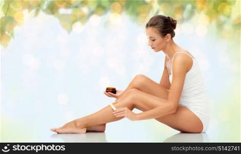 beauty, hair removal and people concept - beautiful woman with applicator applying depilatory wax to her leg over natural green background and lights. beautiful woman applying depilatory wax to her leg