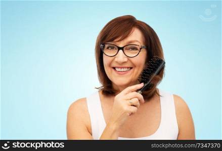 beauty, haircare and old people concept - portrait of smiling senior woman in glasses brushing hair by hairbrush over blue background. portrait of senior woman in glasses brushing hair